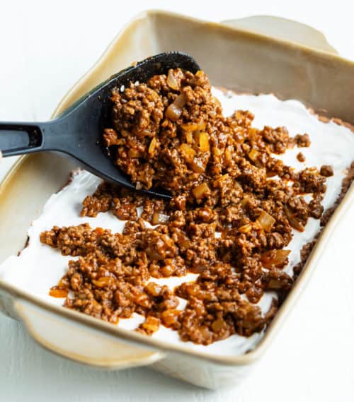 This Taco Casserole is easy to make ahead of time and bake later! It's loaded with juicy ground beef and your favorite Mexican toppings for a perfect weeknight dinner! #doritos #fritos #recipe #mexicanrecipes #easydinnerideas #familydinner #groundbeefrecipes A big spatula spooning seasoned ground beef into a casserole dish for taco casserole.