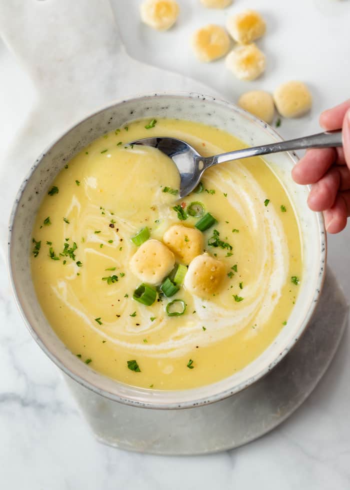 A white bowl with creamy potato leek soup in it with oyster crackers, green onions, and parsley on top.