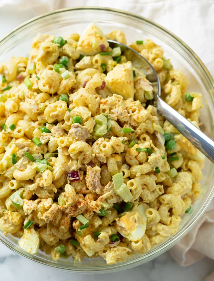 A glass serving bowl filled with Tuna Pasta Salad with a spoon.