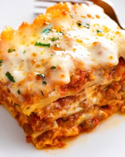 A square slice of cheesy Lasagna with layers of sauce and meat on a white plate.