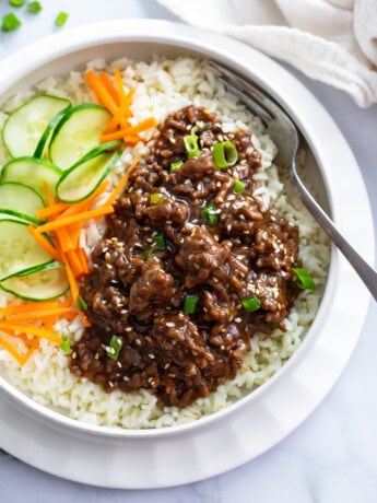 A white bowl of rice topped with Korean ground beef in sauce next to cucumbers and carrots.