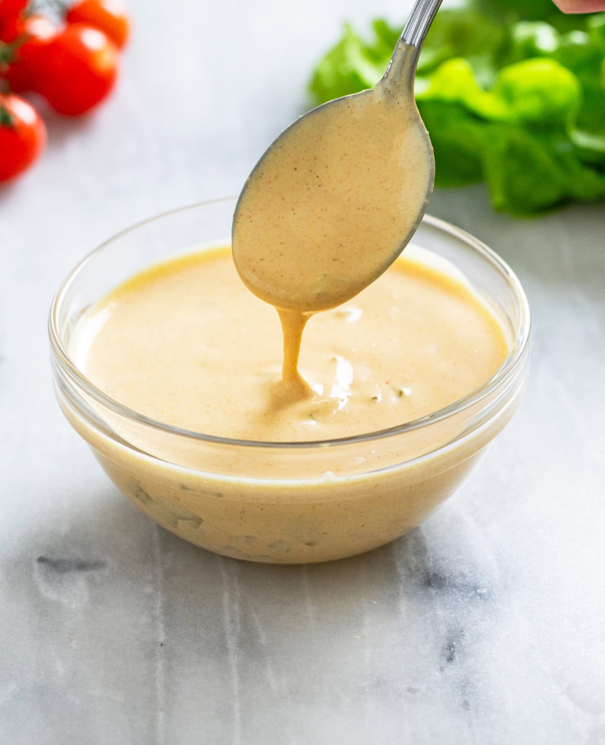 A spoon scooping out Big Mac Sauce from a small glass bowl with lettuce and tomatoes in the background.