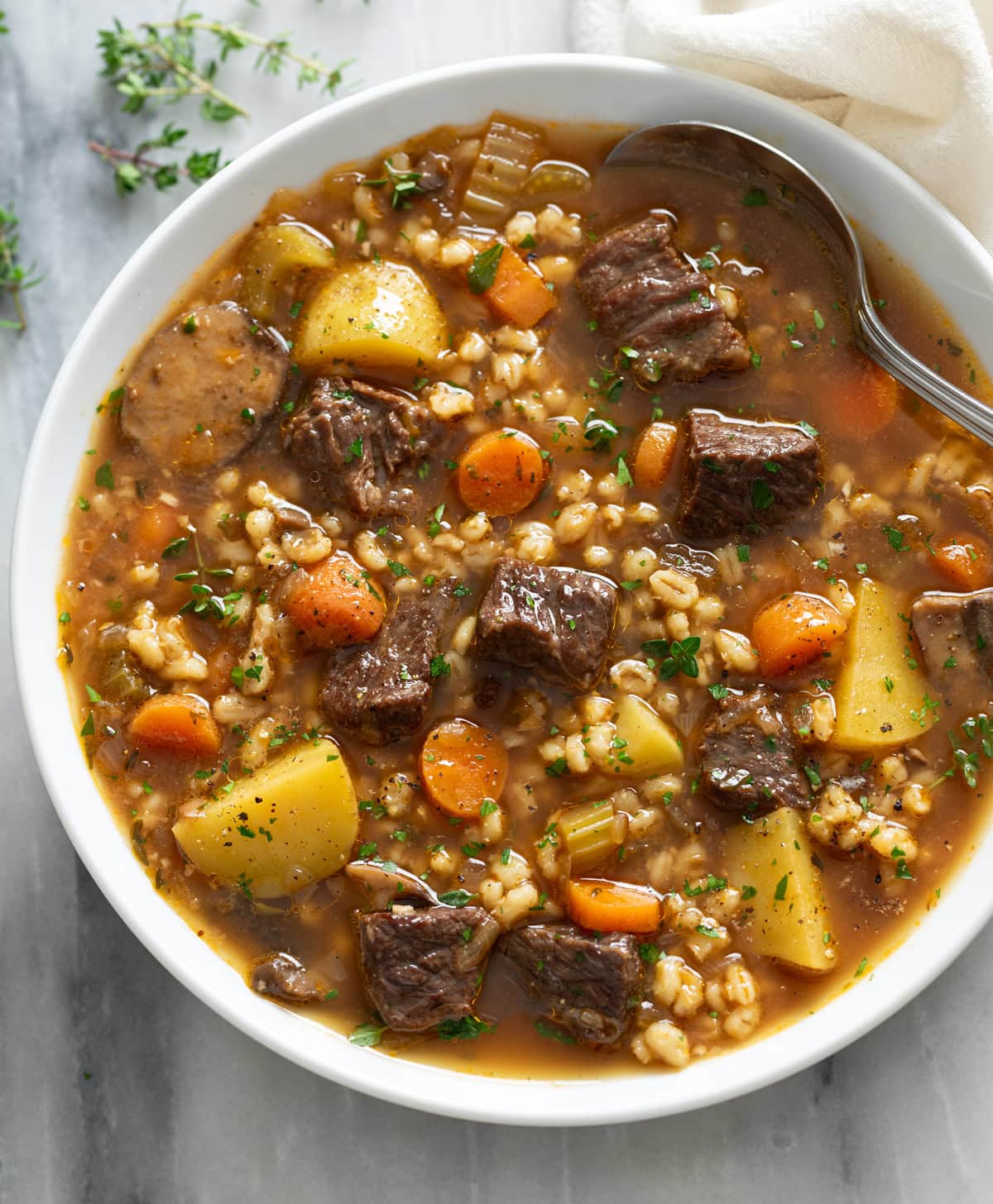 Beef Barley Soup in a white bowl with a spoon on the side and fresh parsley and thyme on top.